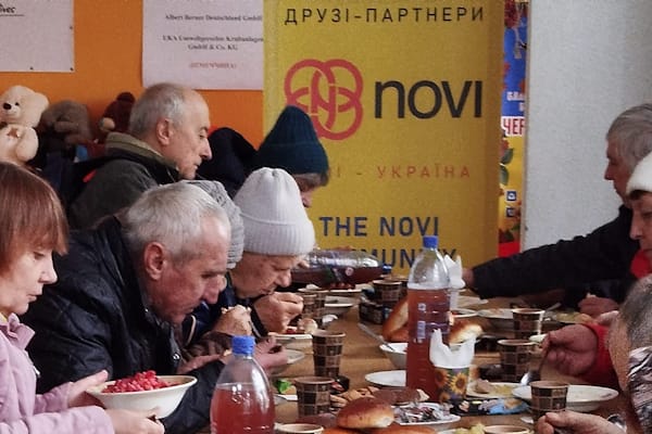 Переселенці за святковим столом насолоджуються обідом, приготованим на Кухні доброти фонду «Червона Калина Харків». Захід організовано за підтримки The Novi Community (США) та ТОВ «ФК “Октава Фінанс” (Україна)».